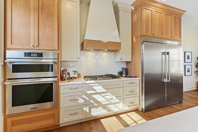 kitchen featuring light countertops, decorative backsplash, light wood-style flooring, appliances with stainless steel finishes, and custom exhaust hood