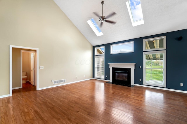 unfurnished living room featuring high vaulted ceiling, a skylight, a fireplace with flush hearth, wood finished floors, and a ceiling fan