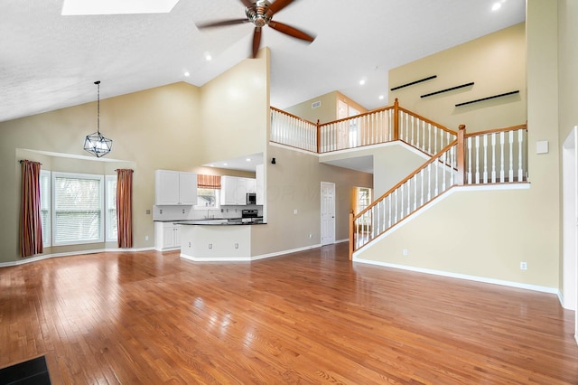 unfurnished living room featuring light wood-style flooring, a ceiling fan, stairs, high vaulted ceiling, and baseboards