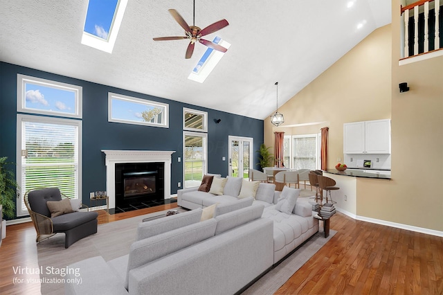 living area featuring high vaulted ceiling, a skylight, ceiling fan, light wood-style flooring, and a tiled fireplace