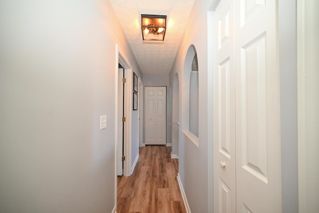 corridor featuring arched walkways, light wood-style flooring, a textured ceiling, and baseboards