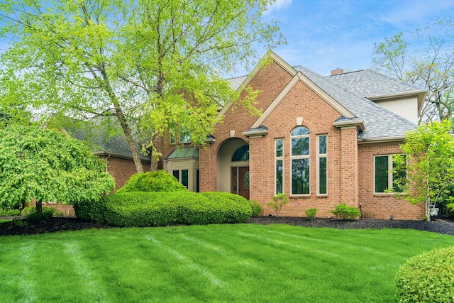 view of front facade featuring a shingled roof, brick siding, a front yard, and a chimney