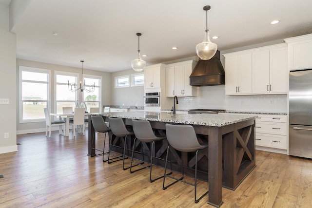 kitchen with custom exhaust hood, stainless steel appliances, a kitchen island with sink, light wood-style floors, and decorative backsplash