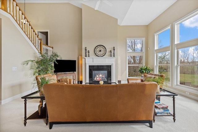 carpeted living area featuring ceiling fan, high vaulted ceiling, a glass covered fireplace, stairway, and baseboards
