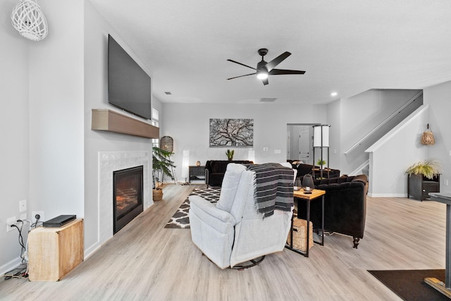 living room featuring light wood finished floors, baseboards, stairs, a ceiling fan, and a glass covered fireplace