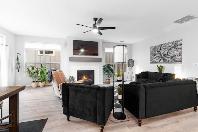 living room featuring a ceiling fan, a warm lit fireplace, light wood finished floors, and visible vents
