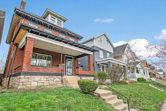 american foursquare style home with covered porch, brick siding, a front yard, and a chimney
