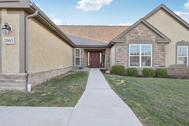 view of exterior entry featuring stucco siding, stone siding, roof with shingles, and a yard