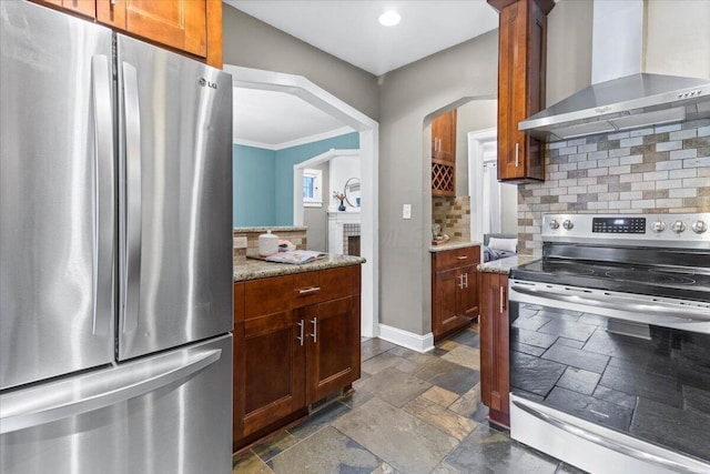 kitchen featuring wall chimney range hood, appliances with stainless steel finishes, arched walkways, decorative backsplash, and stone tile flooring