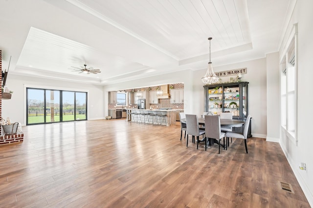 dining space with a raised ceiling, ceiling fan with notable chandelier, wood finished floors, and visible vents