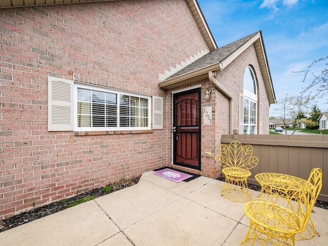 view of exterior entry with brick siding, fence, and a shingled roof