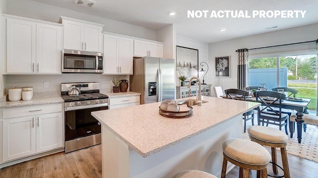 kitchen featuring a kitchen island with sink, light wood-style floors, white cabinetry, stainless steel appliances, and light stone counters