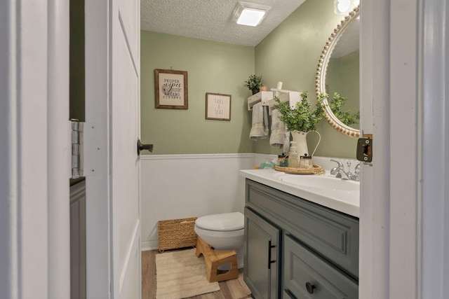 half bathroom with a textured ceiling, wood finished floors, vanity, a wainscoted wall, and toilet