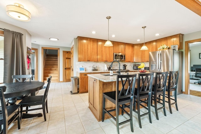 kitchen with stainless steel microwave, light stone counters, a kitchen island with sink, brown cabinetry, and a sink