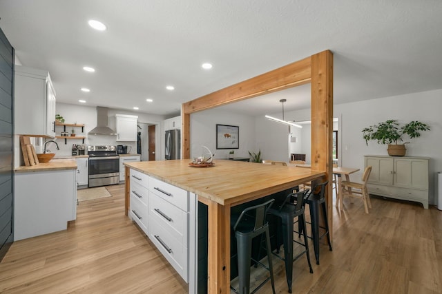 kitchen with wall chimney range hood, open shelves, butcher block counters, a breakfast bar area, and appliances with stainless steel finishes