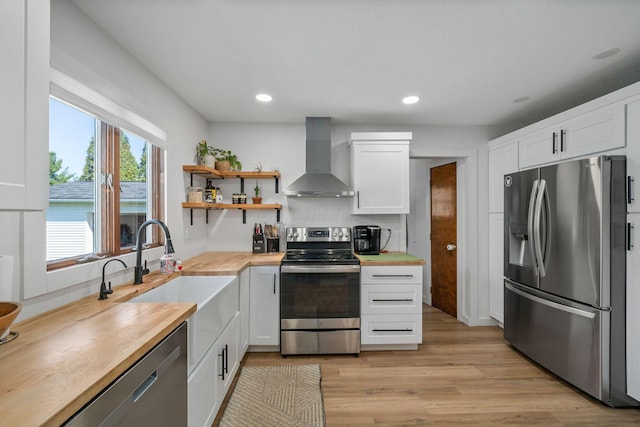 kitchen with light wood-type flooring, wall chimney range hood, appliances with stainless steel finishes, a sink, and wooden counters