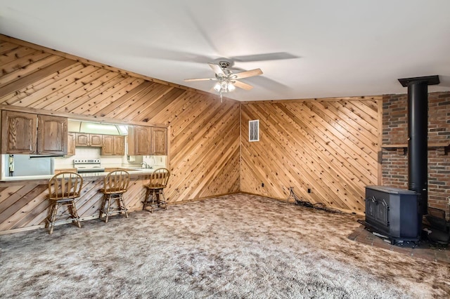 unfurnished living room featuring carpet flooring, ceiling fan, wooden walls, and a wood stove