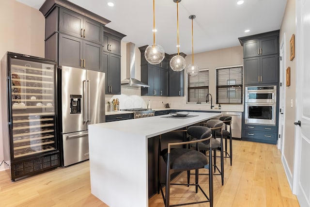 kitchen with wall chimney exhaust hood, light wood finished floors, a sink, beverage cooler, and appliances with stainless steel finishes