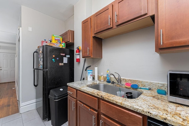 kitchen featuring baseboards, a sink, brown cabinetry, black appliances, and light stone counters