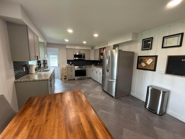 kitchen featuring light stone counters, a sink, stainless steel appliances, backsplash, and gray cabinetry