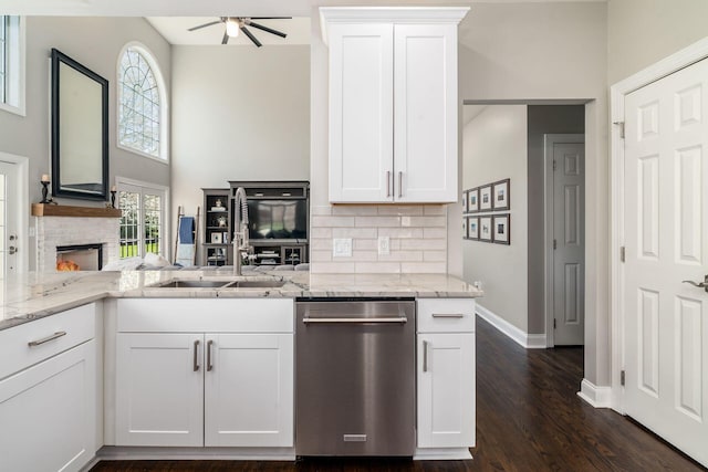 kitchen with a ceiling fan, white cabinets, a stone fireplace, light stone counters, and dishwasher