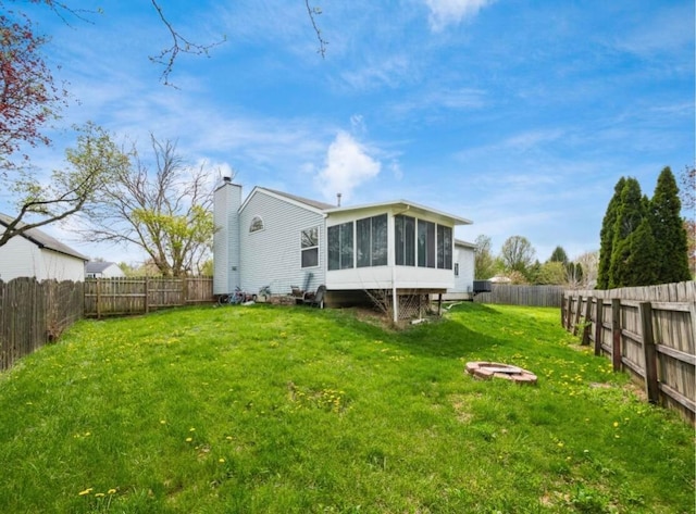 back of house featuring a fire pit, a fenced backyard, a chimney, a sunroom, and a lawn