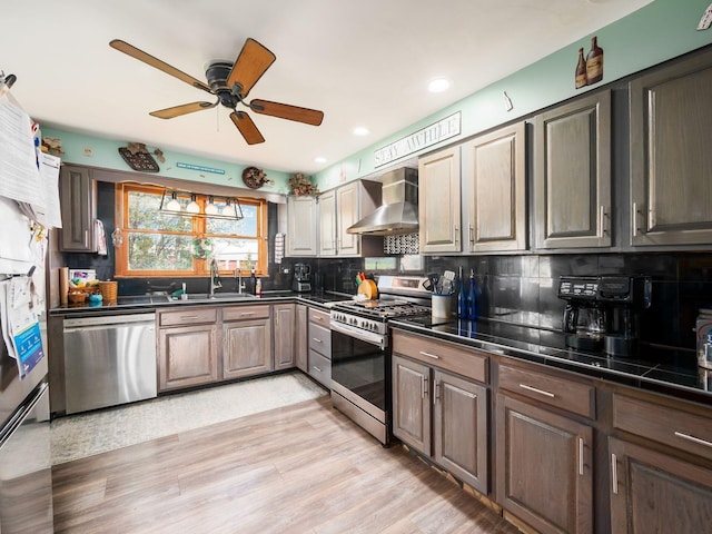 kitchen with a ceiling fan, a sink, wall chimney exhaust hood, dark countertops, and stainless steel appliances