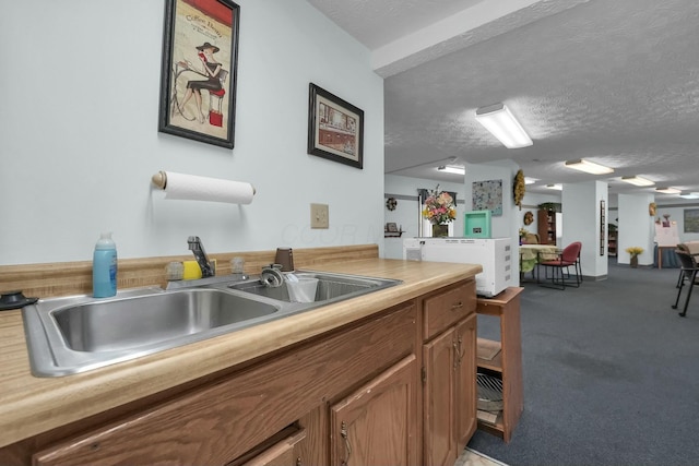 kitchen featuring light countertops, a textured ceiling, brown cabinets, light colored carpet, and a sink