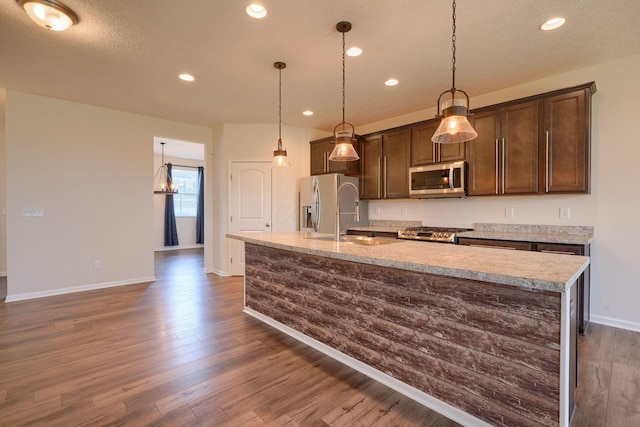 kitchen with dark wood-type flooring, recessed lighting, appliances with stainless steel finishes, a center island with sink, and baseboards
