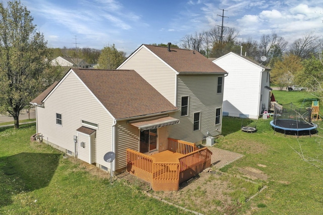 rear view of house featuring a trampoline, a yard, a wooden deck, roof with shingles, and central AC