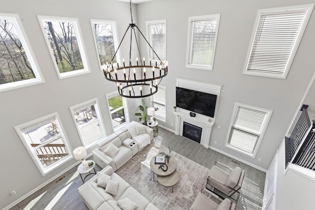 living room featuring baseboards, a notable chandelier, wood finished floors, a towering ceiling, and a glass covered fireplace
