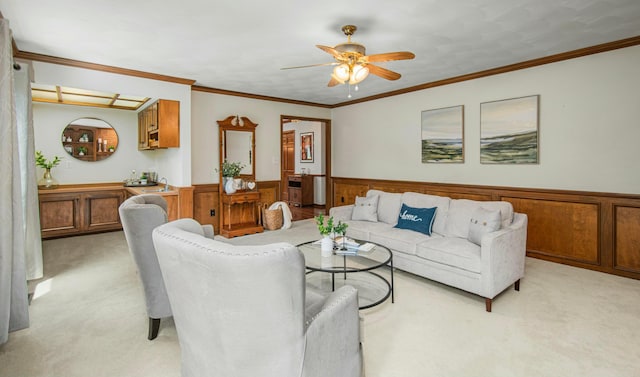 living room with crown molding, a wainscoted wall, a ceiling fan, and light carpet