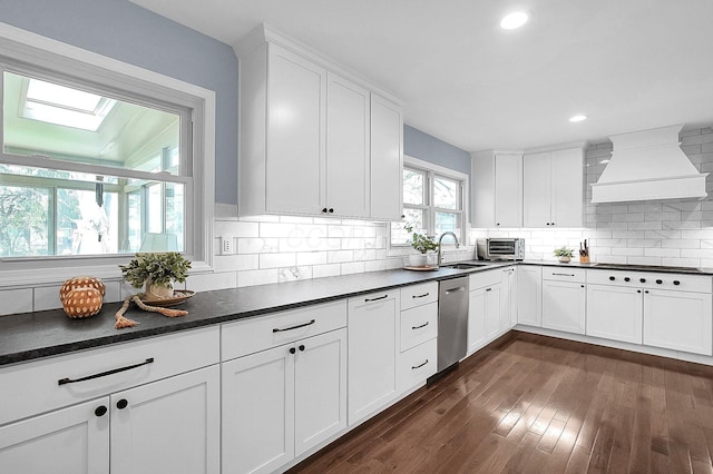 kitchen featuring a skylight, dark countertops, premium range hood, stainless steel dishwasher, and decorative backsplash