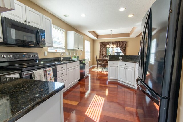 kitchen with a sink, black appliances, a tray ceiling, white cabinetry, and dark wood-style flooring