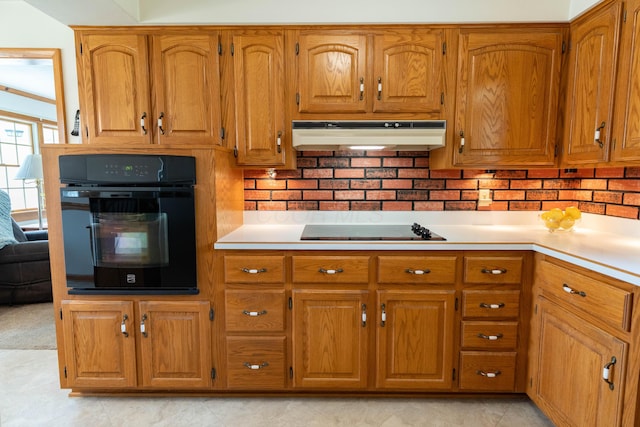 kitchen featuring brown cabinetry, black appliances, under cabinet range hood, light countertops, and decorative backsplash