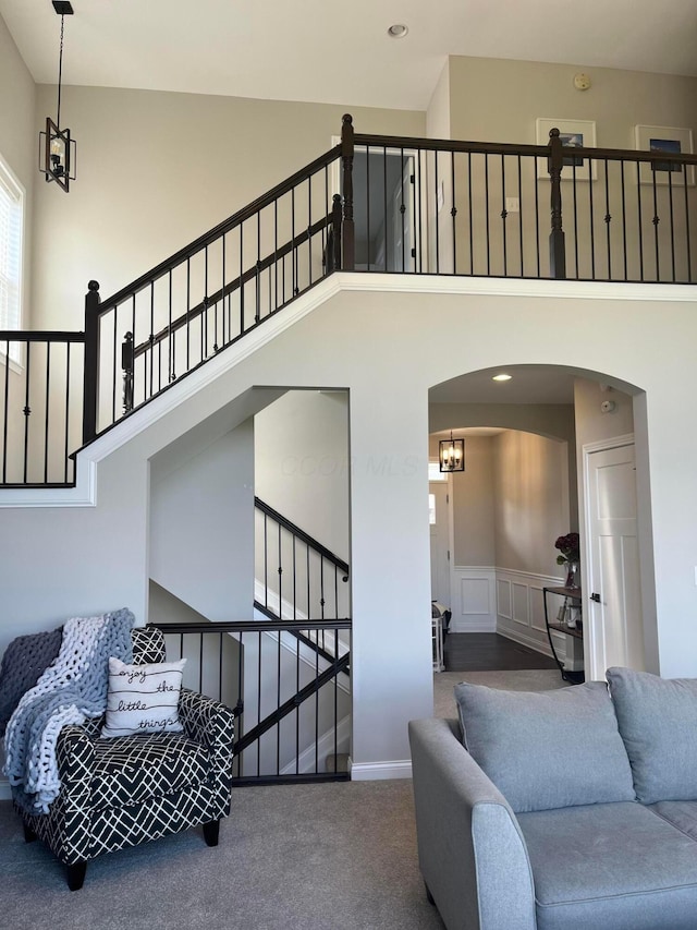 carpeted living area featuring arched walkways, a high ceiling, a notable chandelier, and wainscoting