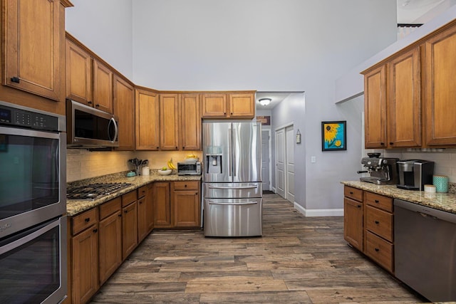 kitchen with tasteful backsplash, a towering ceiling, brown cabinets, dark wood-type flooring, and appliances with stainless steel finishes