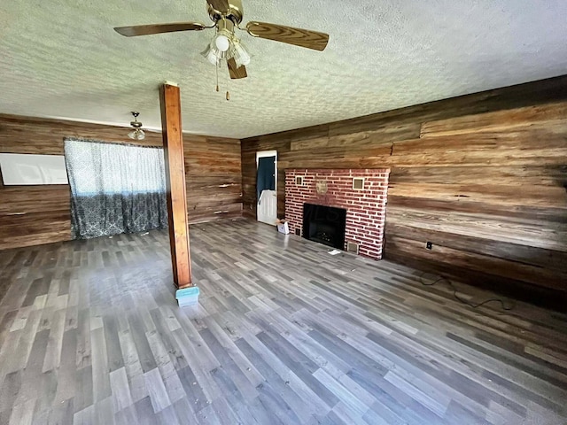 unfurnished living room with a textured ceiling, wood walls, wood finished floors, a brick fireplace, and ceiling fan