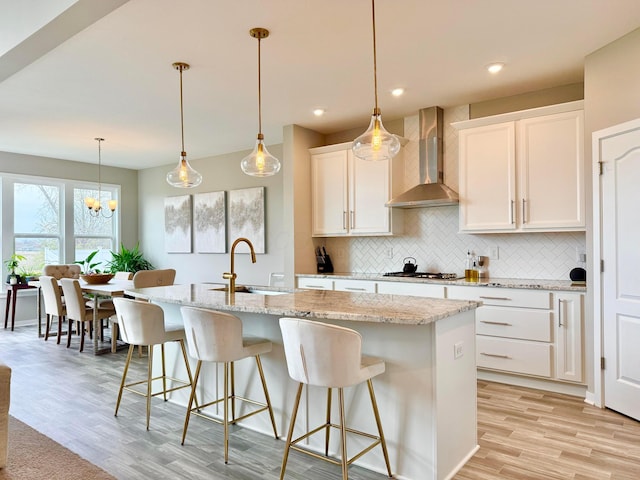 kitchen featuring wall chimney range hood, a sink, light wood finished floors, a center island with sink, and backsplash