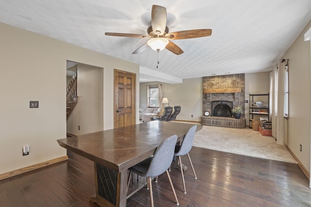 dining space with stairway, ceiling fan, baseboards, a brick fireplace, and dark wood-style floors