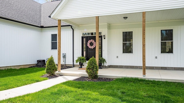 view of exterior entry with a yard, roof with shingles, and covered porch