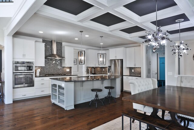 kitchen featuring wall chimney range hood, a chandelier, stainless steel appliances, dark wood-style flooring, and a center island with sink