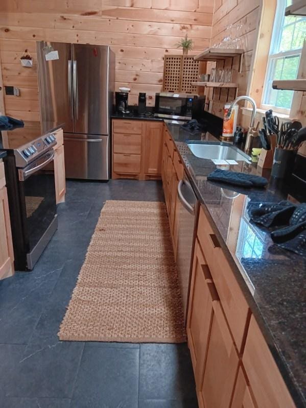 kitchen featuring light brown cabinets, wooden walls, a sink, dark stone countertops, and appliances with stainless steel finishes