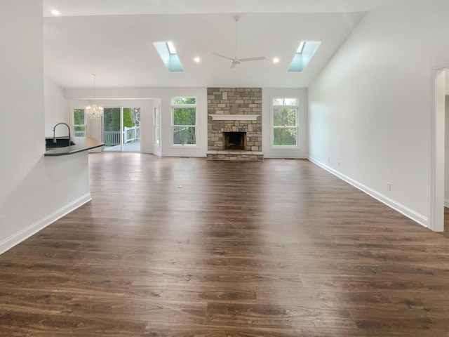 unfurnished living room featuring lofted ceiling with skylight, a stone fireplace, ceiling fan with notable chandelier, baseboards, and dark wood-style floors