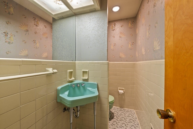 half bath with toilet, tile walls, wallpapered walls, a wainscoted wall, and a sink