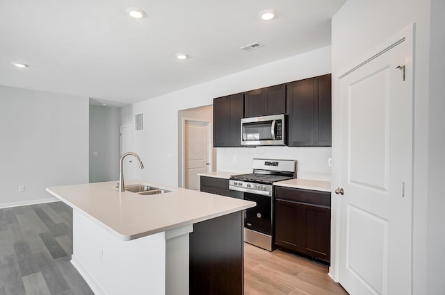 kitchen with light wood finished floors, light countertops, a sink, a kitchen island with sink, and appliances with stainless steel finishes