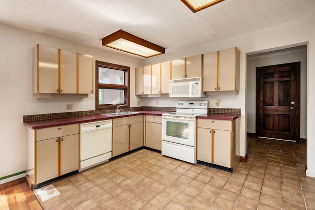 kitchen with a textured ceiling, a sink, dark countertops, cream cabinetry, and white appliances