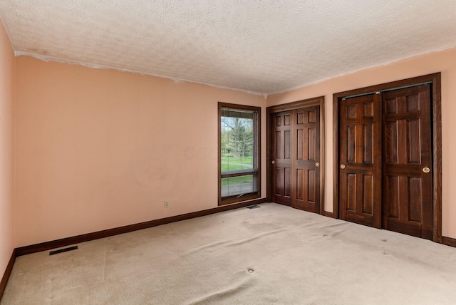 unfurnished bedroom with a textured ceiling, light colored carpet, visible vents, baseboards, and two closets