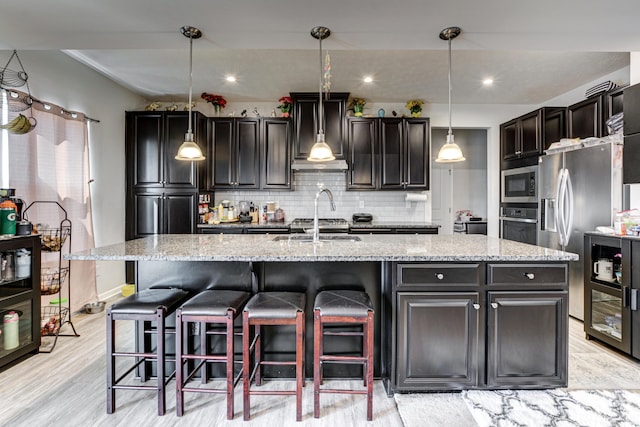 kitchen with a center island with sink, light wood-type flooring, tasteful backsplash, light stone counters, and appliances with stainless steel finishes