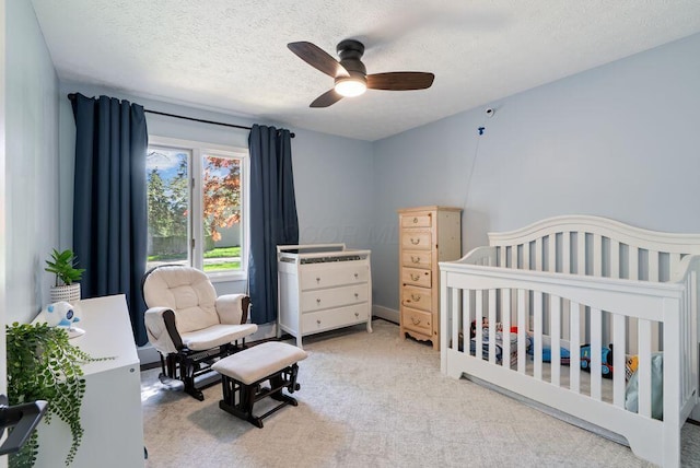 bedroom with a textured ceiling, light colored carpet, a crib, and ceiling fan